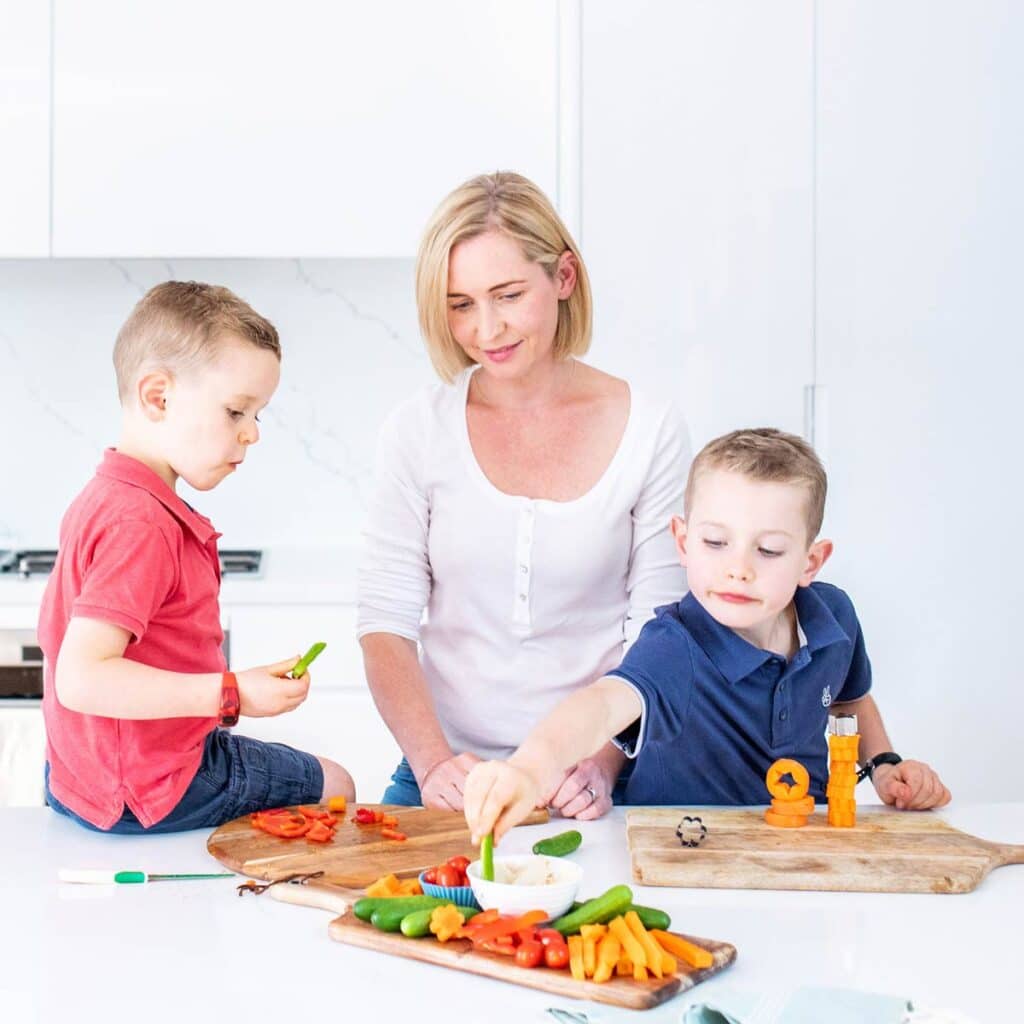 Amy in the kitchen with her two sons preparing and eating finger vegetables on wooden trays.