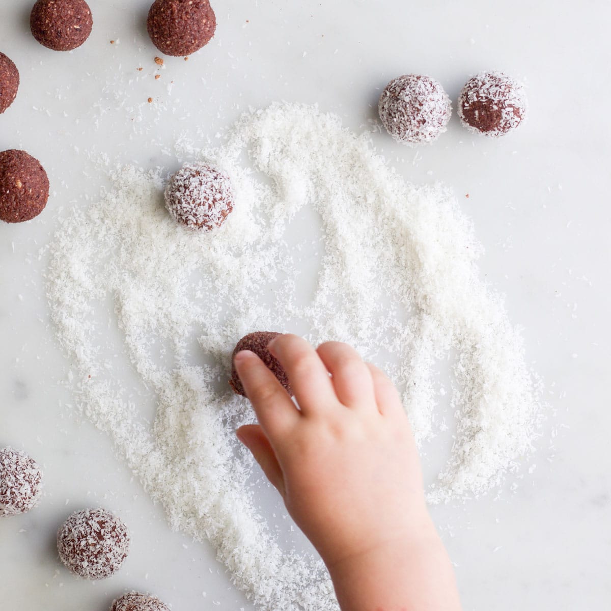 Child's Hand Rolling Cherry Ball in Coconut with Coated and Uncoated Balls in Background.