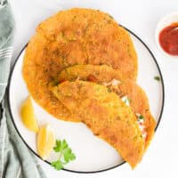 White Plate With Two Large Chickpea Pancakes On. One Folded Over. Lemon on the Plate and Green Cloth in Background.