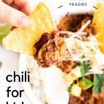 Child's Hand Holding Tortilla Chip Topped with Chilli With Bowl of Chilli in Background. Text overlay "chili for kids 5 Hidden Veg"