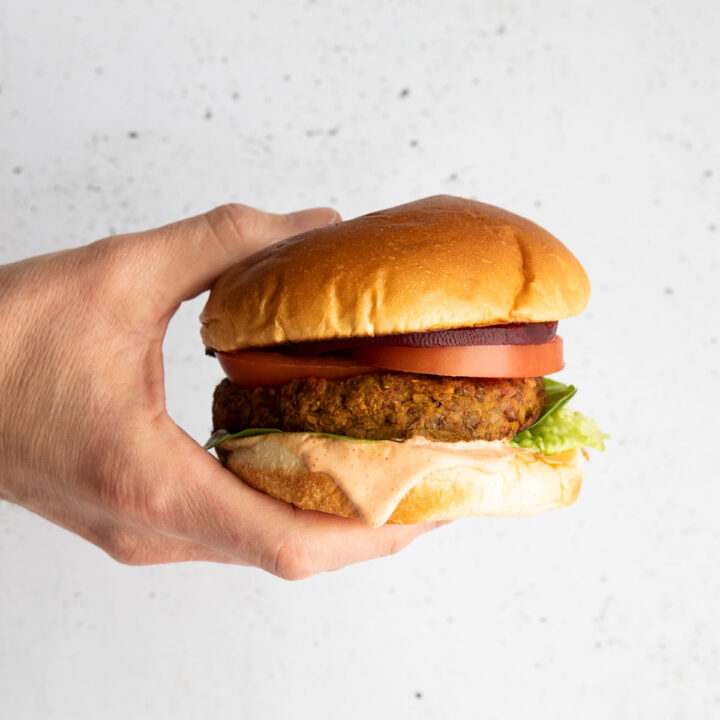 Hand Holding Lentil Burger in Bun with Burger Sauce, Lettuce, Tomato and Beetroot.