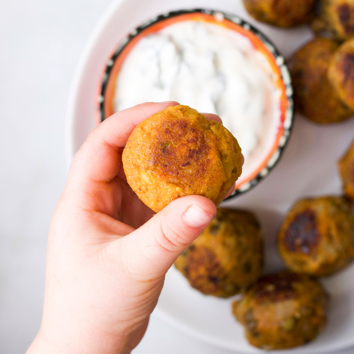 Child's Hand Holding a Lentil Burger Ball With Other Balls and Dip in Background.