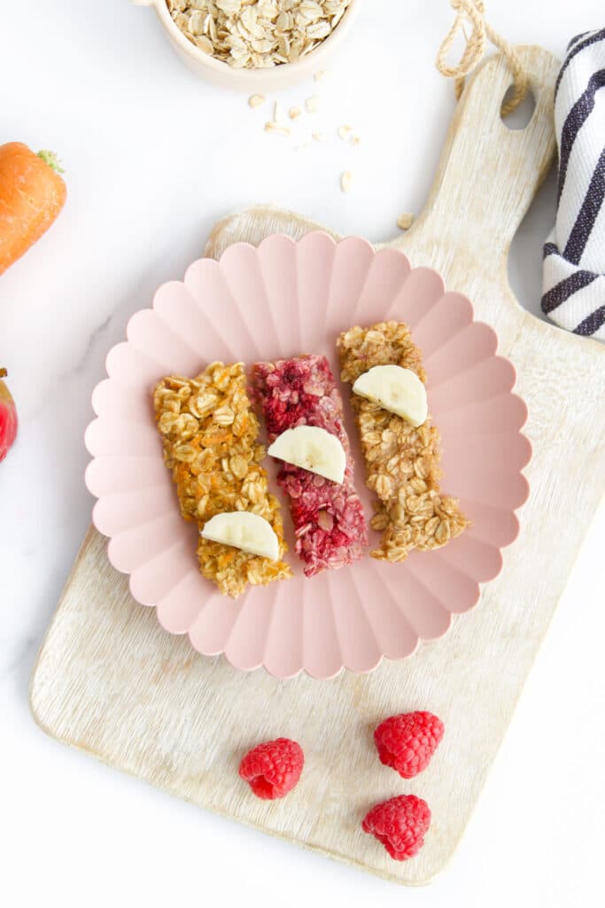 3 Porridge Fingers on Flower Shaped Plate Sitting on Board with Raspberries, Carrot and Oats in Background.