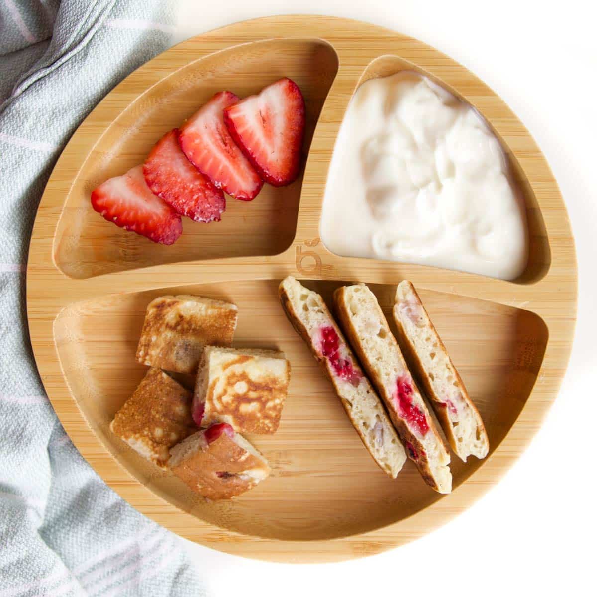 Divided Wooden Baby Plate with Strawberry Pancakes Cut into Fingers and Bite Sized Pices. Sliced Strawberries and Yogurt in Other Sections