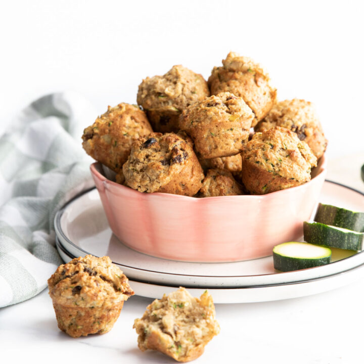 Mini Zucchini Muffins IN Bowl with Sliced Zucchini and Half Eaten Muffin in Background.